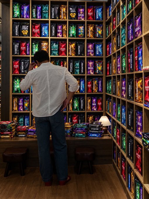 A man in a white shirt and jeans stands looking at a wall filled with neatly organized shelves containing various brightly colored snack packages like 'STARTER', 'GRAL', and 'ELITE'.