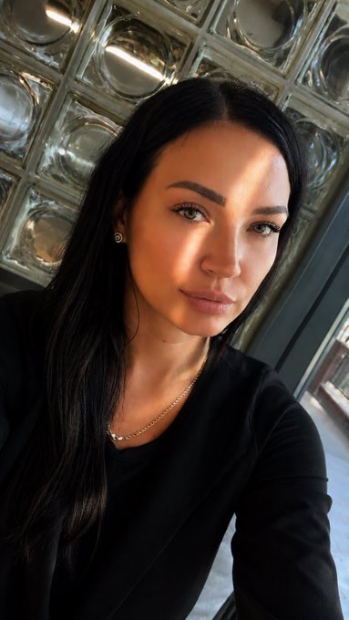 A close-up portrait of a young woman with long dark hair wearing a black blazer and a delicate silver necklace, illuminated by warm sunlight against a glass block wall.