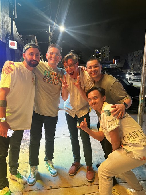 A group of five friends pose together on a nighttime city street, with one wearing a BAYC-themed graphic tee, smiling and flashing peace signs as they celebrate their meetup ahead of ApeFest being held at Beeple's studios.