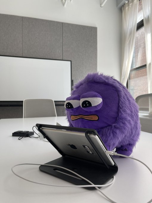 A fuzzy purple plush toy wearing a stern face, set up in a meeting room as if it were working on a tablet propped on a stand.