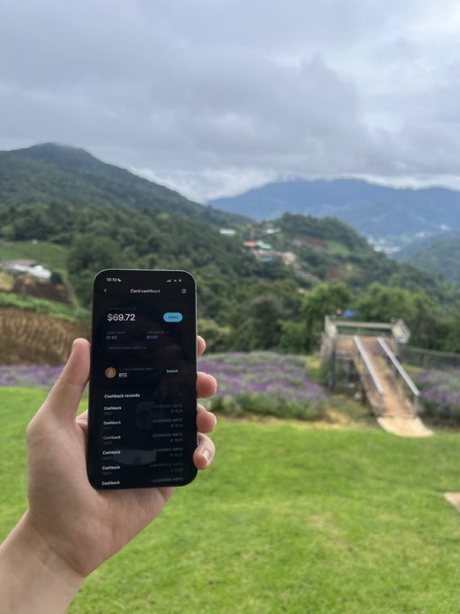 A hand holds a smartphone displaying a "Card cashback" app screen showing a $69.72 balance, set against a scenic backdrop of mountains, greenery, and purple flowers, visually tying the idea of earning crypto rewards through AssetBack on Bitget while enjoying life's beautiful moments.