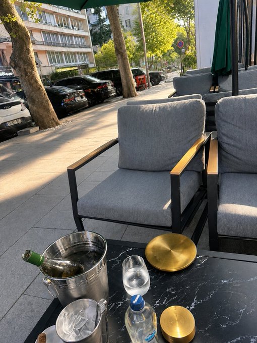An outdoor café patio featuring gray cushioned chairs around a black marble table with a wine bucket, glasses, and water bottles.