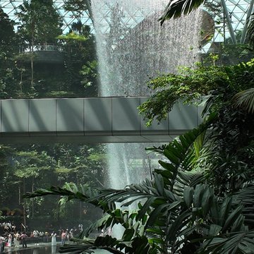 An indoor view of a massive waterfall cascading from a glass ceiling, surrounded by lush tropical greenery with a walkway bridge crossing in front of it.