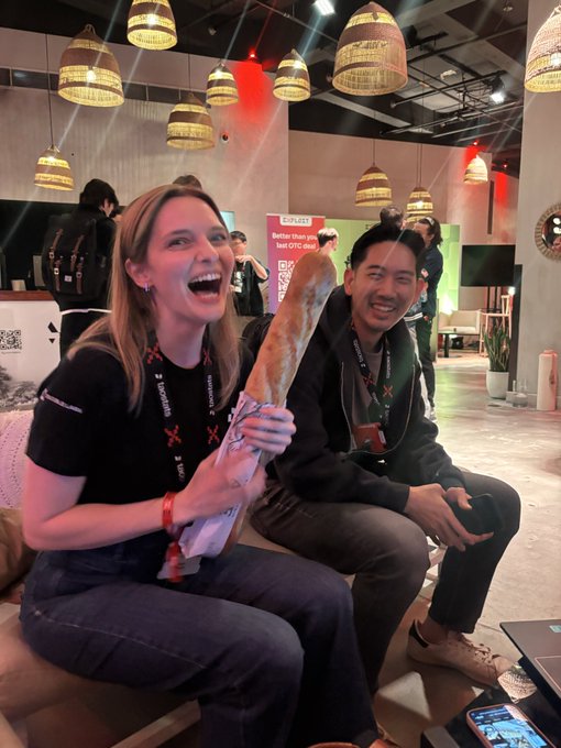 Two smiling people wearing lanyards sit at an event; the woman on the left holds up a long baguette enthusiastically.