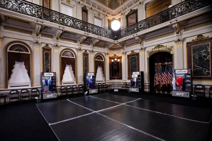 The image shows a large, empty room with a black floor and white walls. The room has several chairs placed around the space, some of which are adorned with posters or banners. There are also two TVs mounted on the wall, one closer to the center of the room and another towards the right side. A clock is visible in the upper part of the image. 

The room appears to be a large, open space designed for various activities such as watching UFC fights or other events. The presence of chairs and posters suggests that this could be an event setup or a gathering area within a venue where people can sit and enjoy the presentations or performances taking place.