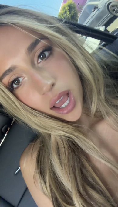 Close-up selfie of a blonde woman with highlighted hair sitting inside a car, wearing a black top.