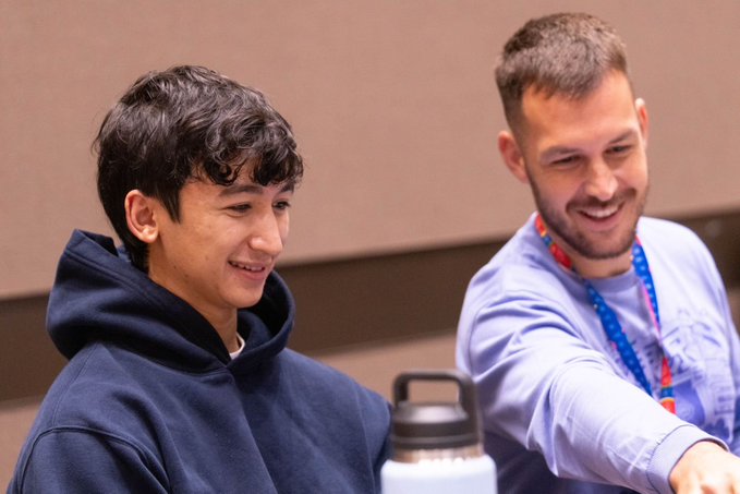 Two men sit side by side smiling warmly, with the younger one in a dark hoodie on the left and an older bearded man in a light blue shirt with a lanyard on the right. A white water bottle sits on the table.