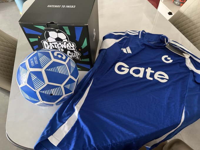 The image features a blue soccer jersey with white stripes, placed next to a soccer ball and a box of soccer balls. The soccer ball has a unique design resembling a blue and white hexagon. The scene takes place around a dining table where chairs are positioned on both sides. A person can be seen in the background, possibly preparing for or enjoying a meal while surrounded by these items.