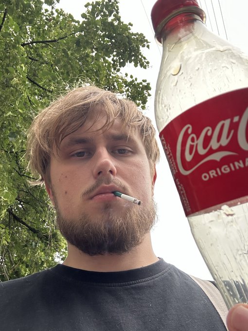 A selfie-style photo of a young bearded man with a cigarette in his mouth, holding up a large Coca-Cola bottle outdoors with trees in the background.