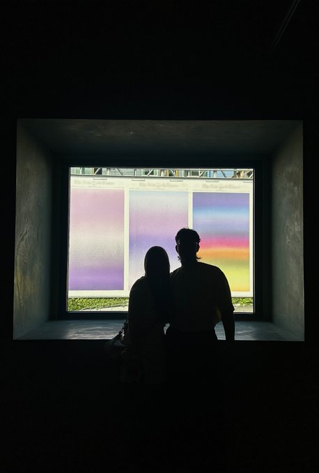 A silhouette of two people sitting together in a dark room, framed by a bright window with three colored glass panels in purple, white, and a sunset gradient.