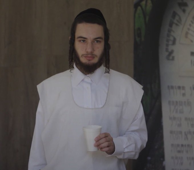 A young man wearing a yarmulke and white ceremonial garments with a bib, holding a paper cup next to a Hebrew-inscribed gravestone.
