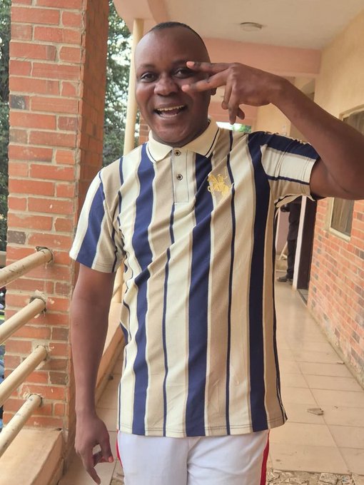 A smiling man in a navy-and-cream striped polo poses with a hand sign on a brick-column veranda.