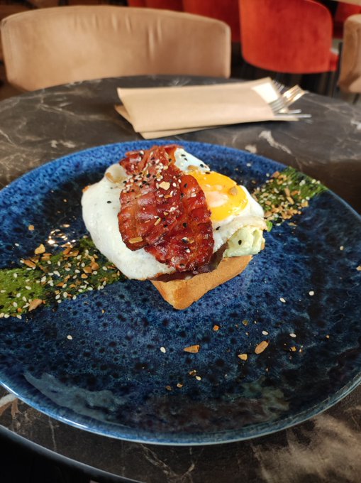 A fried egg and bacon served on toast with avocado, garnished with furikake, on a blue plate.