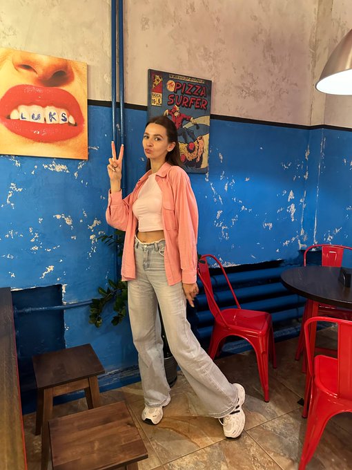 A woman in a pink shirt and light blue jeans posing with a peace sign in a colorful cafe with blue walls, red chairs, and pop art paintings including a 'PIZZA SURFER' poster and a lips artwork.