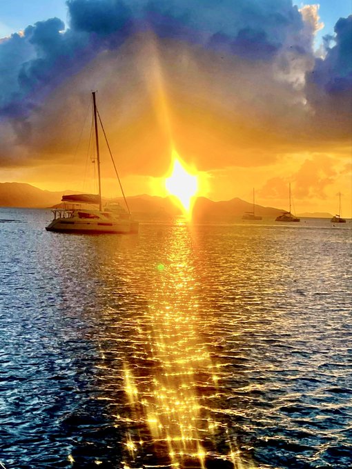 A scenic sunset over the ocean with a catamaran and other boats silhouetted against the golden sun reflecting on the water.