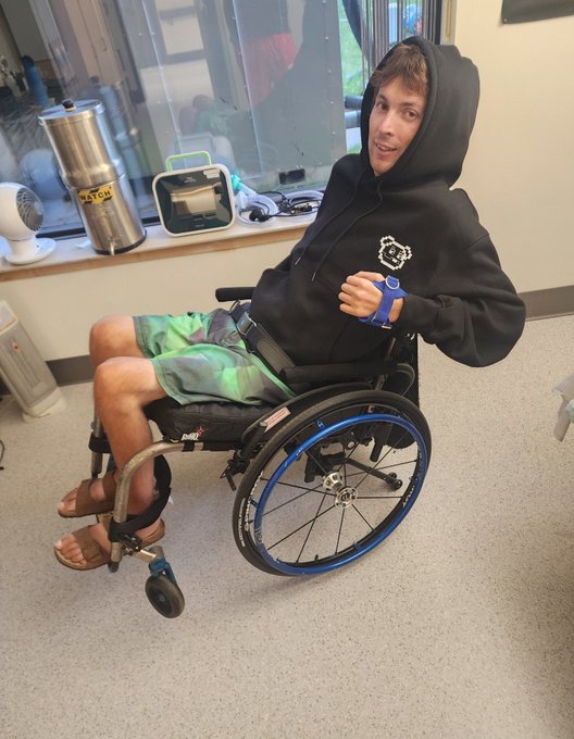 A young man in a black hoodie and green shorts sitting in a wheelchair inside a medical facility, smiling and wearing a blue wrist brace.