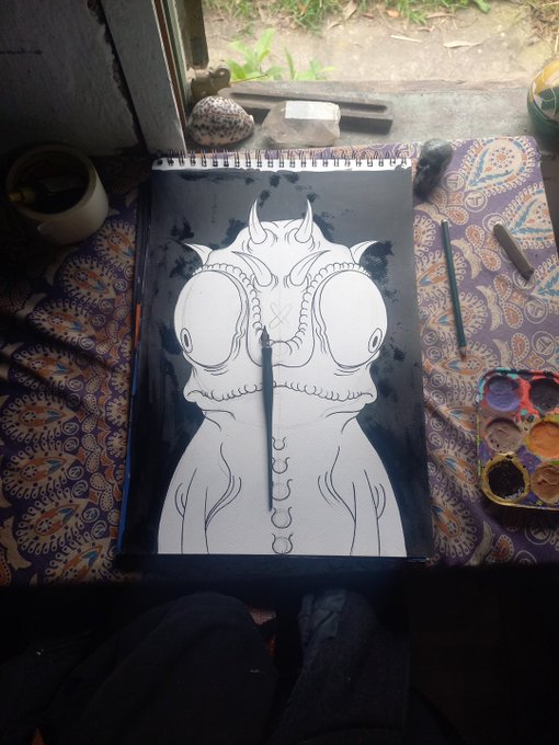 An open sketchbook on a patterned cloth displays a black ink drawing of a horned creature, accompanied by a paintbrush, pencils, a seashell, and a paint palette.
