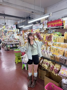 A young woman poses for a photo in what appears to be a snack aisle of a Thai market or convenience store, surrounded by colorful packaged goods and signs in Thai. The image accompanies a post about stepping away from her full-time crypto role, skipping Token2049, and vacationing while attending The Weeknd concert in Thailand.