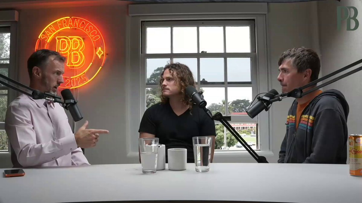 The image features three men sitting at a table, with two of them holding microphones while one man holds a can of soda. The background includes a window that allows natural light into the room. A neon sign reading "bitcoin" is visible on the wall behind the men. The text in the image suggests that Bitcoin has been stolen and is being used to create fake currency called Cuckoo coins, which are printed from thin air and can't be redeemed due to their inherent vulnerability.
