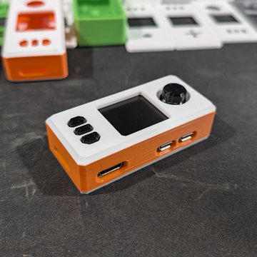 A white and orange handheld gaming device or electronic gadget with a small screen, buttons, and USB ports, resting on a dark surface with similar devices blurred in the background.