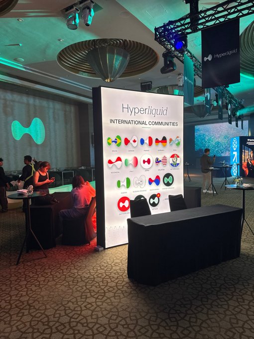 An event photo showing a 'Hyperliquid INTERNATIONAL COMMUNITIES' display board with various country flags, set up at what appears to be a conference with green-lit ceiling decor.