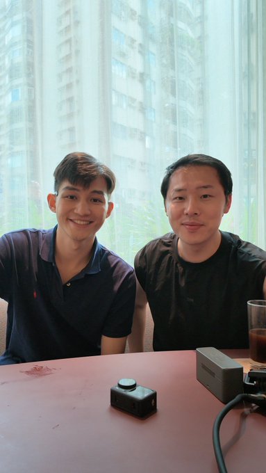 Two men sitting together at a pink table smiling for a photo, with two small camera devices placed in front of them and a tall building visible through the window behind.
