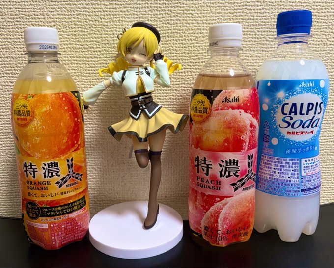 A yellow-haired anime figure stands on a white base between three beverages: an Asahi '特濃' (Tokunou) Orange Squash, an Asahi '特濃' Peach Squash, and a Calpis Soda bottle.