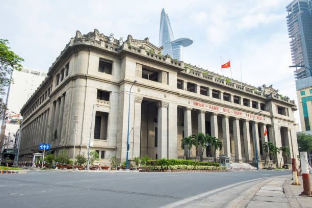 A large, classical-style stone building with columns and the text 'NGÂN HÀNG NHÀ NƯỚC' on its facade, with a Vietnamese flag and the Bitexco Financial Tower skyscraper visible in the background.