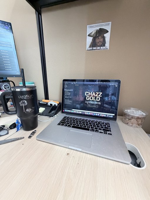 A desk setup featuring a laptop displaying 'CHAZZ GOLD', a black 'Ovation' tumbler, glasses, and a pirate meme poster on the wall.