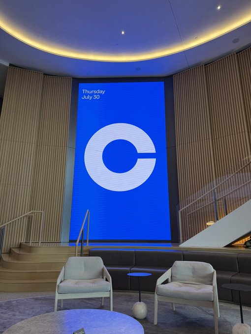A modern lounge area features a large digital screen displaying the Coinbase logo, dated 'Thursday July 30', with light wood paneling and two armchairs.