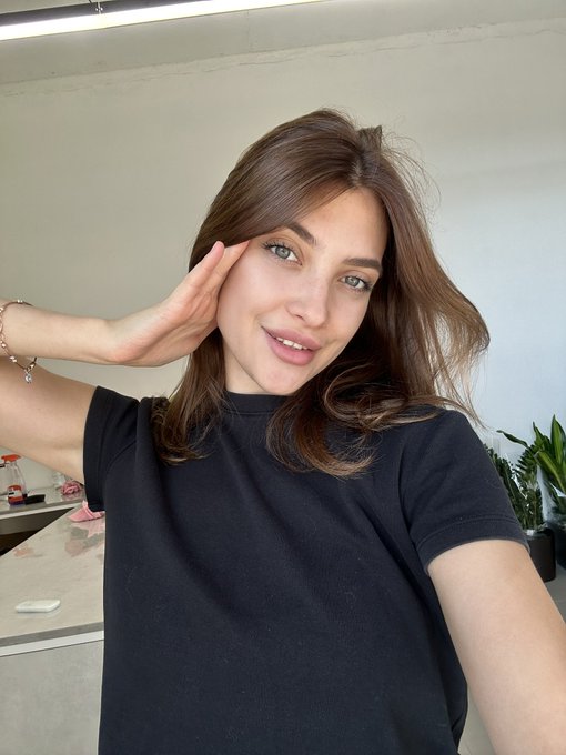 A selfie of a smiling young woman with long brown hair wearing a black t-shirt, posing with her hand near her face in a room with plants.
