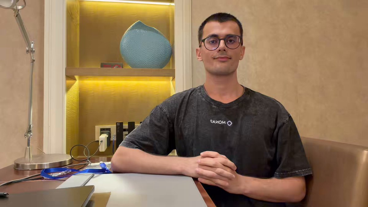 The image features a man sitting at a desk with his hands crossed, wearing glasses and a black t-shirt. The desk has an open laptop computer placed on top of it. In front of him, there are two books - one blue and one white. A vase is also present in the scene, located near the center of the image. The man appears to be posing for a photo or participating in a video call, possibly related to the post "monad foundry day 2 - the basics of @monad".