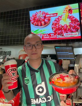 A man wearing glasses and a green and black striped jersey holds a cup of coffee and a bowl of food inside a restaurant, with a digital menu board showing seafood dishes in the background.