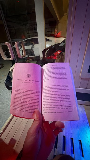 A first-person view of someone reading an open paperback book titled 'THE GOLDEN RULE OF HABIT CHANGE' in a dimly lit area, with another person visible in the background.