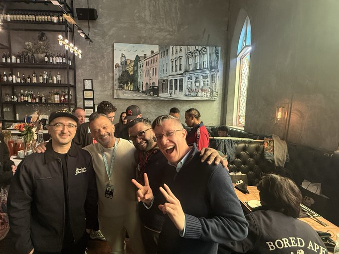 A group of men posing happily for a photo at a bar or pub, with one making a peace sign, set against a backdrop of liquor shelves and a stained glass window; a person in the foreground wears a jacket reading 'BORED APES'.