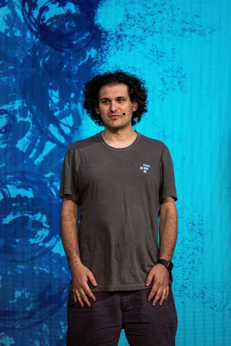 A young man with curly hair wearing a dark gray FTX t-shirt stands in front of a large blue screen, appearing to speak at an event.