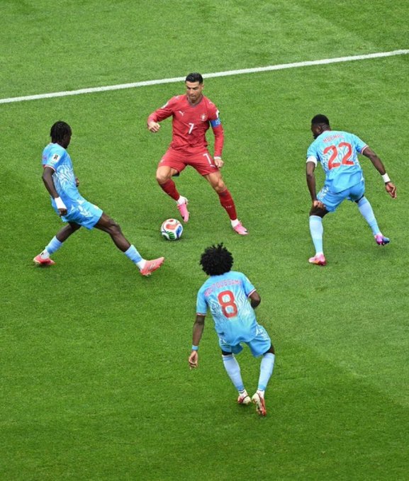 The image features a group of soccer players dressed in blue uniforms, with one player wearing a red jersey. They are playing a game on a grassy field, and there's a sports ball visible near them. The main focus of the image is the soccer ball, which appears to be the center of attention for the players as they engage in their match.
