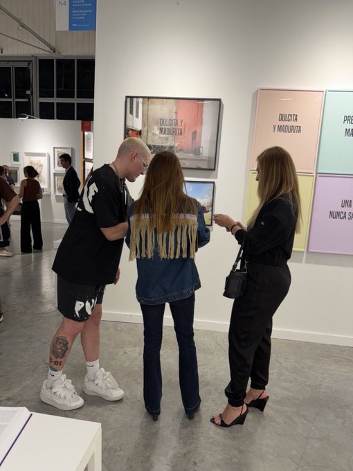 Three people view artwork at an art gallery, with framed pieces displaying Spanish text including 'DULCITA Y MAUDUITA' and 'UNA NUNCA SA' on the walls.