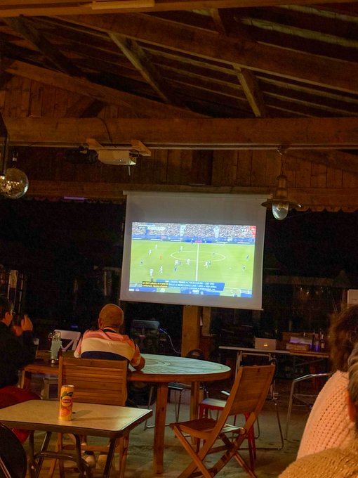 People are watching a soccer match projected on a screen inside a rustic, barn-like venue with exposed wooden beams and ceiling fans.