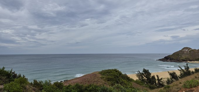 A coastal landscape view shows a calm blue ocean under a cloudy sky, with a sandy beach and green hills on the right side.
