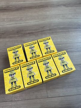 The image features a collection of six BNB binance boxes, all of which are yellow with black text that reads "BNB" or "binance", placed on a wooden floor. The boxes appear to be identical, suggesting they might be part of a promotional campaign for Binance, the cryptocurrency and financial services company founded by Brian Winton in 2013.