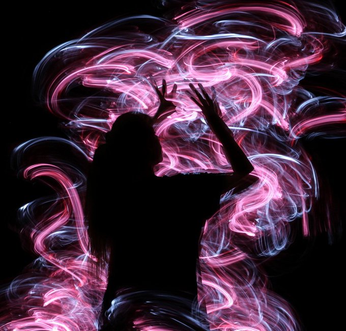 The image features a person with long hair standing in front of an abstract background, possibly resembling watercolors or neon art due to its colorful and ethereal appearance. The individual appears to be captivated by the light painting artwork they are viewing, as their pose suggests curiosity and excitement towards this unique form of visual expression.