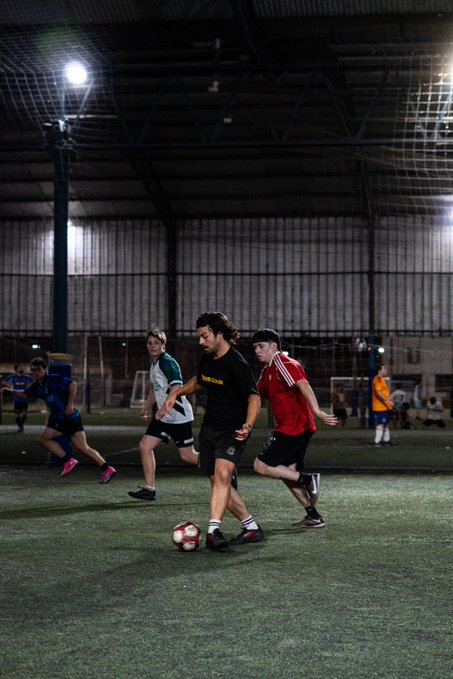 Young men playing soccer on an indoor turf field at night under bright lights, with one player in a black jersey about to kick the ball.