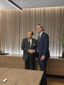 Two men in business suits posing together and shaking hands in what appears to be a corporate meeting room setting.