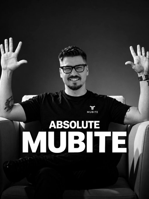 A black-and-white promotional image features a smiling man with glasses, a beard, and tattoos seated in a chair with both hands raised in an enthusiastic gesture, wearing a "MUBITE" branded t-shirt. Bold overlay text reads "ABSOLUTE MUBITE," visually reinforcing the post's announcement of major platform upgrades—faster speed, smoother UI, and upcoming big updates for crypto prop trading.