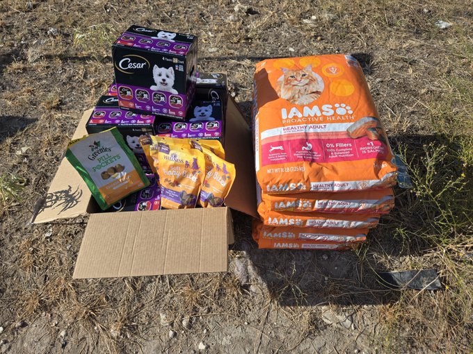 A cardboard box filled with dog supplies, including purple Cesar dog food trays, a bag of Dr. Gary's Best Breed dog treats, and a stack of orange IAMS ProActive Health dog food bags, sits on dry grass awaiting distribution. The image reflects the post's message of community-driven, real-world impact by showcasing tangible goods being prepared for donation or delivery through SP3ND's offline charitable efforts.