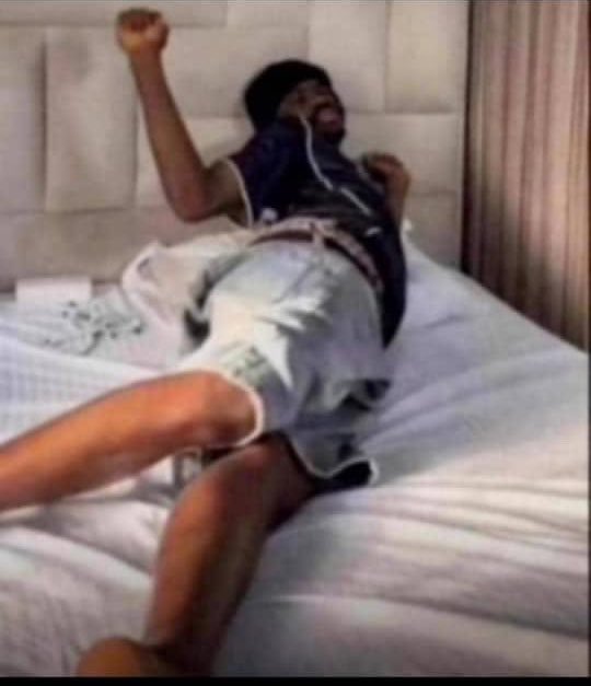 A low-quality photo shows a man wearing a basketball jersey and shorts lying back on a white bed in a celebratory pose with one arm raised and a fist clenched.