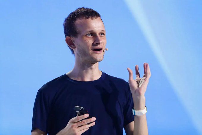 The image features a man wearing a blue shirt, standing on stage with his hands raised as if he's giving a speech or presentation. The background of the image is a light blue color that contrasts with the man's dark blue shirt. There are no other people in the image apart from the speaker and himself.