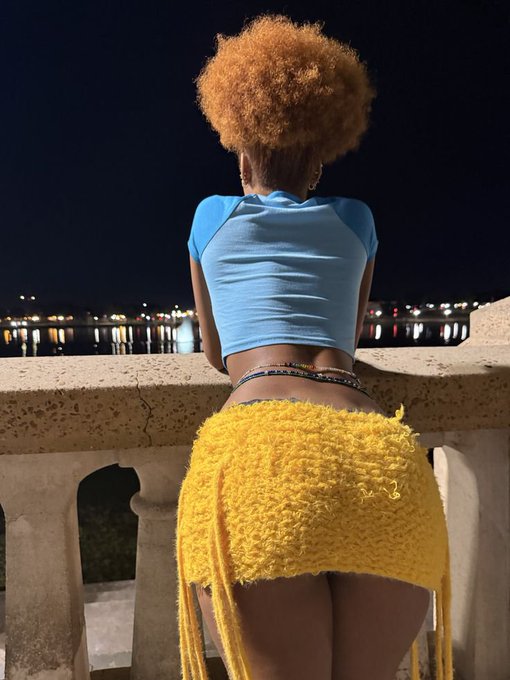 A woman with a high afro hairstyle wearing a yellow knit skirt and blue crop top stands facing away at a nighttime waterfront overlooking a harbor with city lights reflecting across the water.