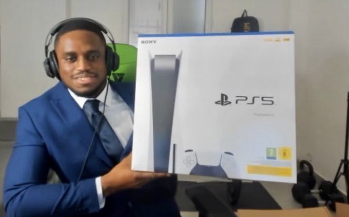 A man in a blue suit and tie wearing headphones smiles while holding a large box for a Sony PlayStation 5 console.